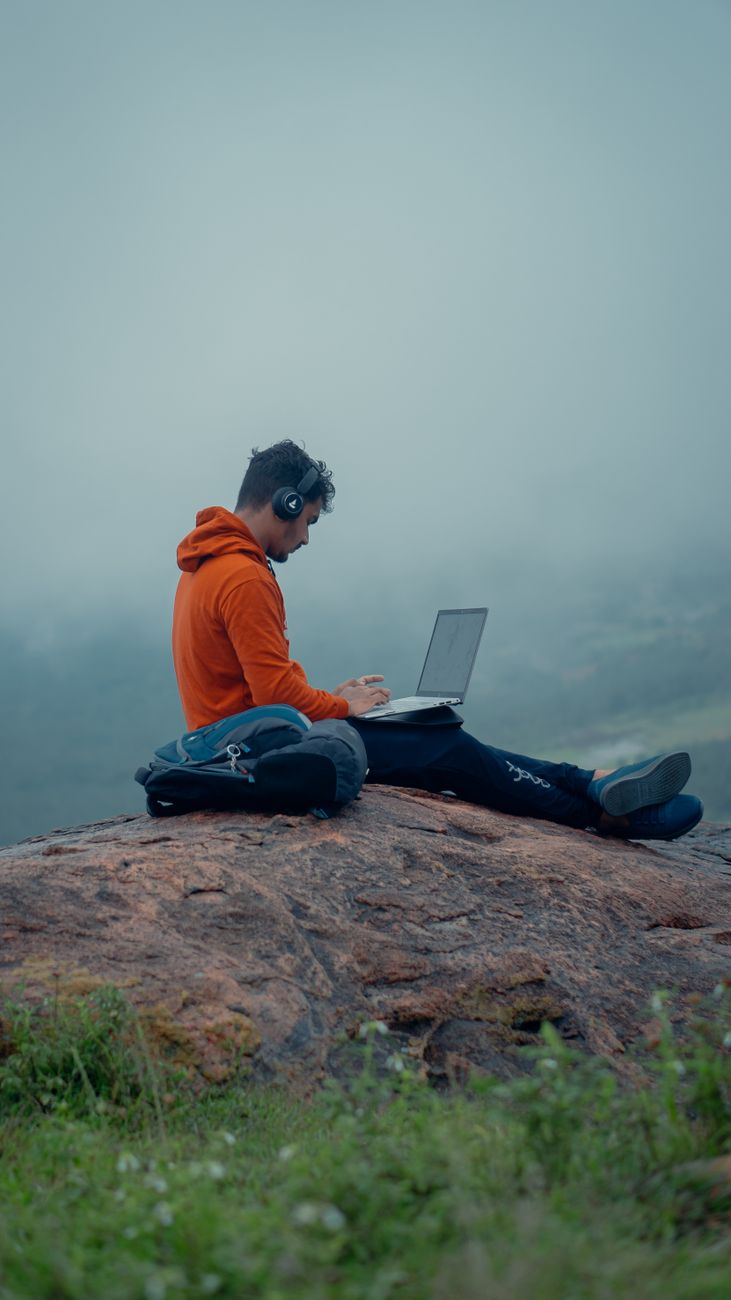 man in orange hoodie sitting on brown rock using laptop